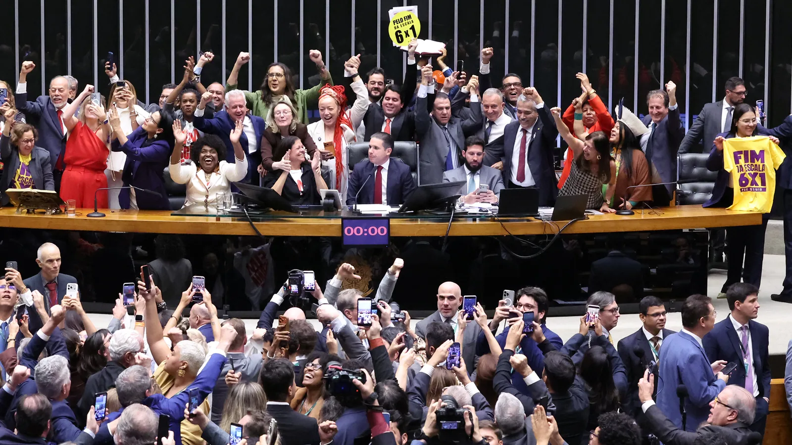Deputados comemoram de braços erguidos na Mesa e no plenário da Câmara a aprovação da PEC que acaba com a escala 6x1; ao fundo, um cartaz amarelo com 6x1