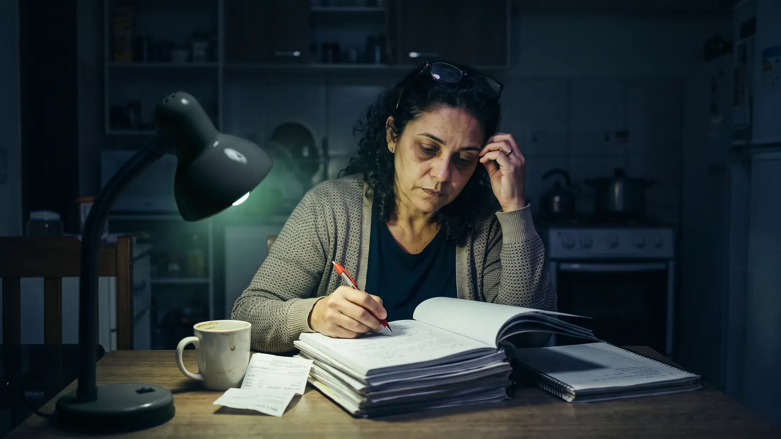 Professora cansada corrige uma pilha de cadernos à noite na mesa da cozinha, sob a luz verde de um abajur